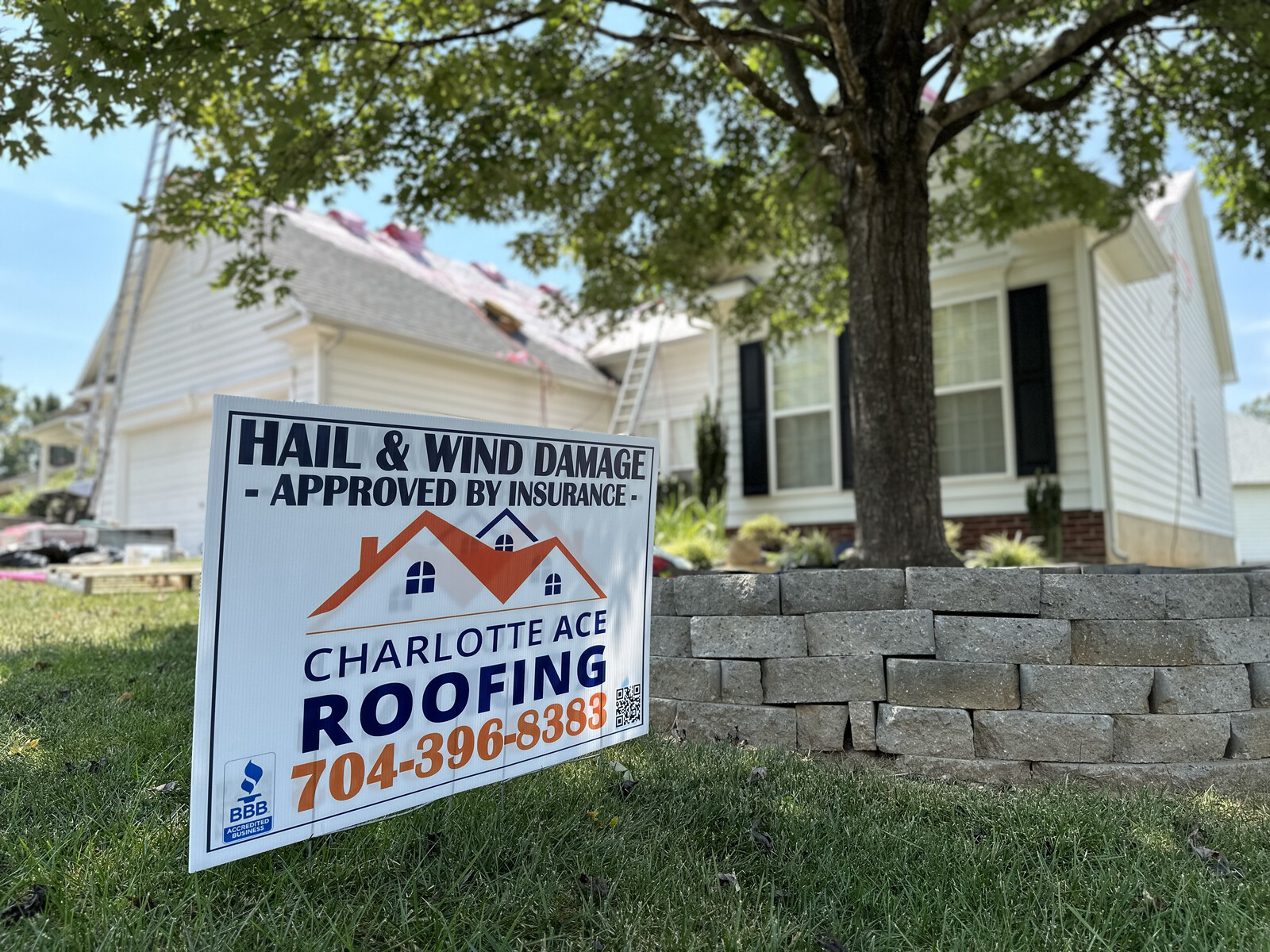 Roof replacement in progress on a Belmont, NC home \u2014 Charlotte Ace Roofing yard sign with BBB Accredited Business badge in foreground, crew installing new shingles in background