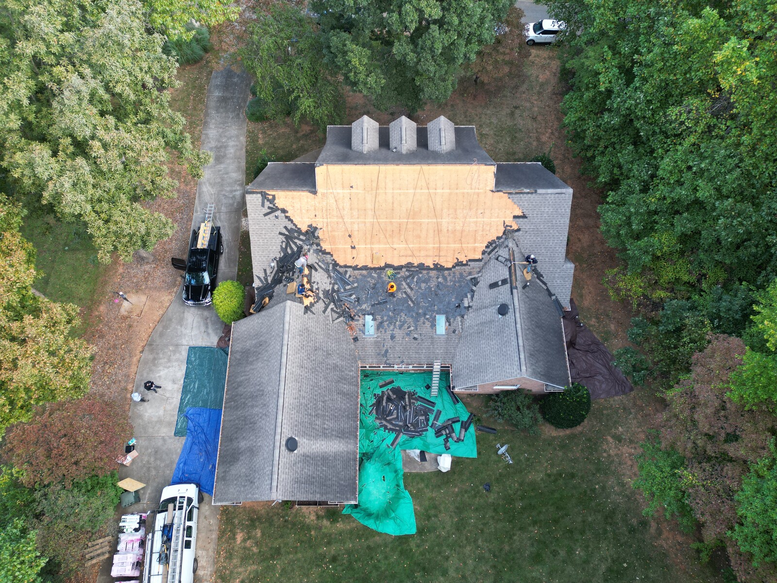 Aerial drone view of a Belmont, NC roof tear-off in progress \u2014 Charlotte Ace Roofing crew on the deck, white company van and dump truck in driveway, large two-story home in wooded lot
