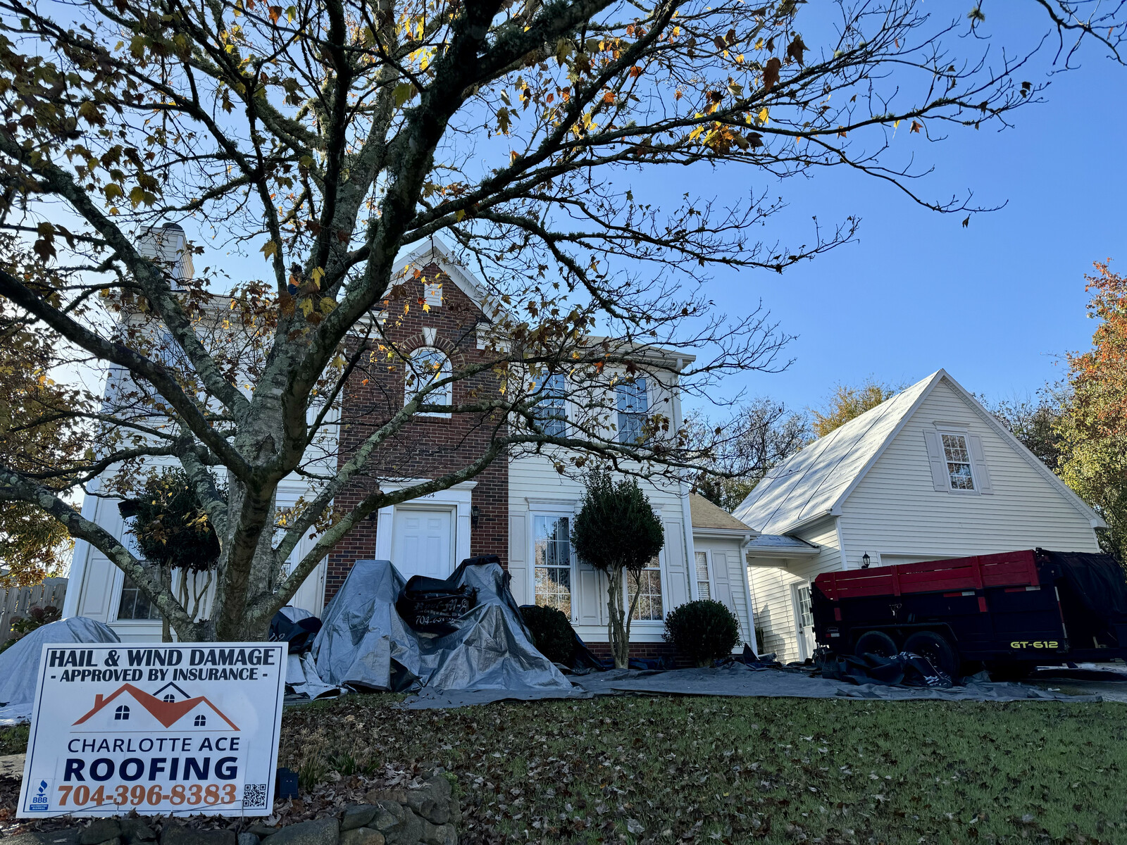 Active roof tear-off project at a brick traditional home in East Charlotte &mdash; tarps protecting landscaping, dump trailer in driveway, Charlotte Ace Roofing yard sign