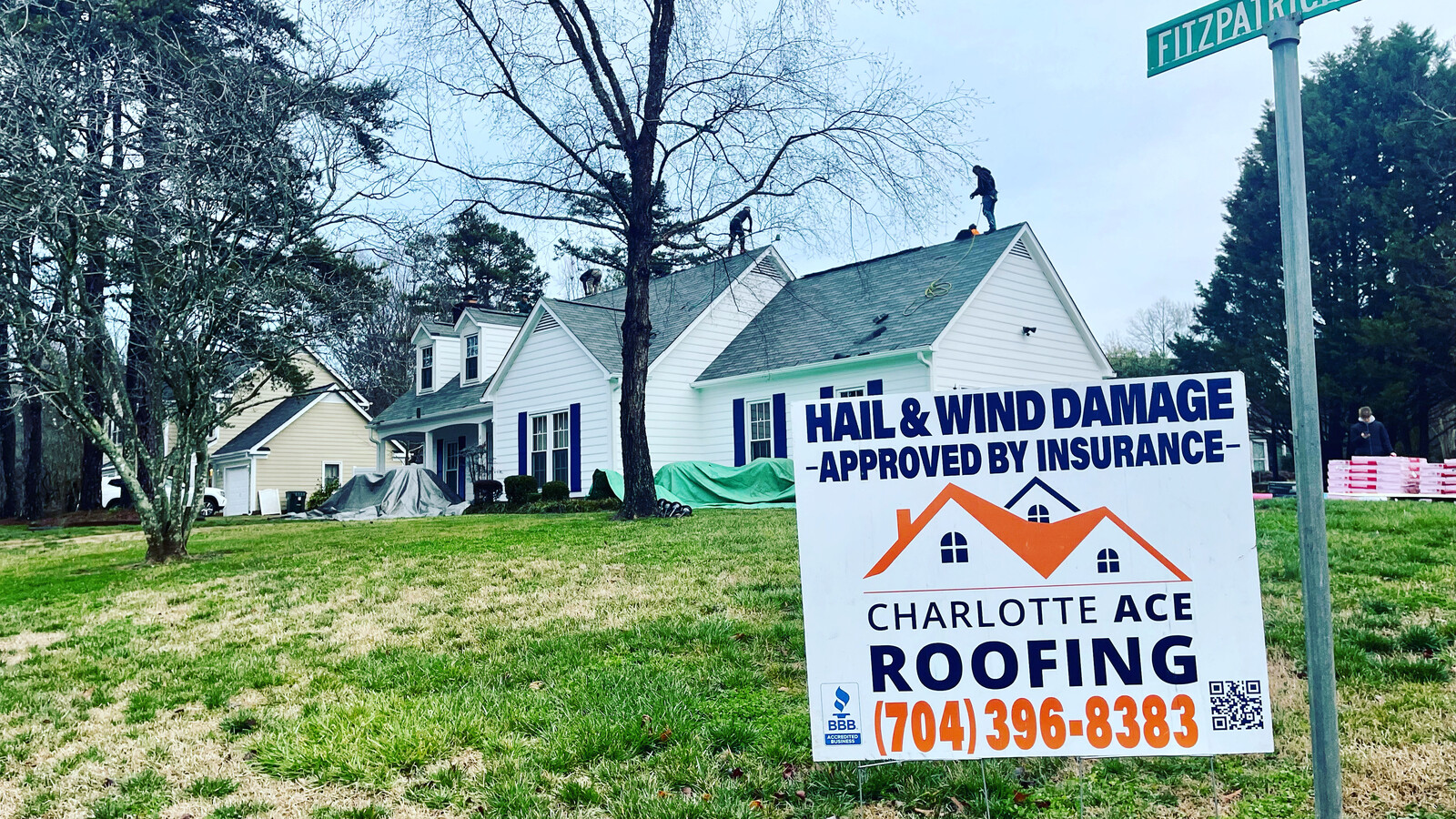 Active Charlotte Ace Roofing crew installing a new roof on a traditional white home with navy shutters &mdash; IKO shingle bundles staged in the driveway, yard sign in foreground