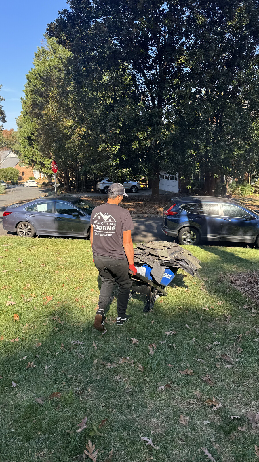 Charlotte Ace Roofing crew member in branded shirt hauling old shingle tear-off debris in a Cobalt wheelbarrow across a Highland Creek-area Charlotte NC residential lawn during a roof replacement
