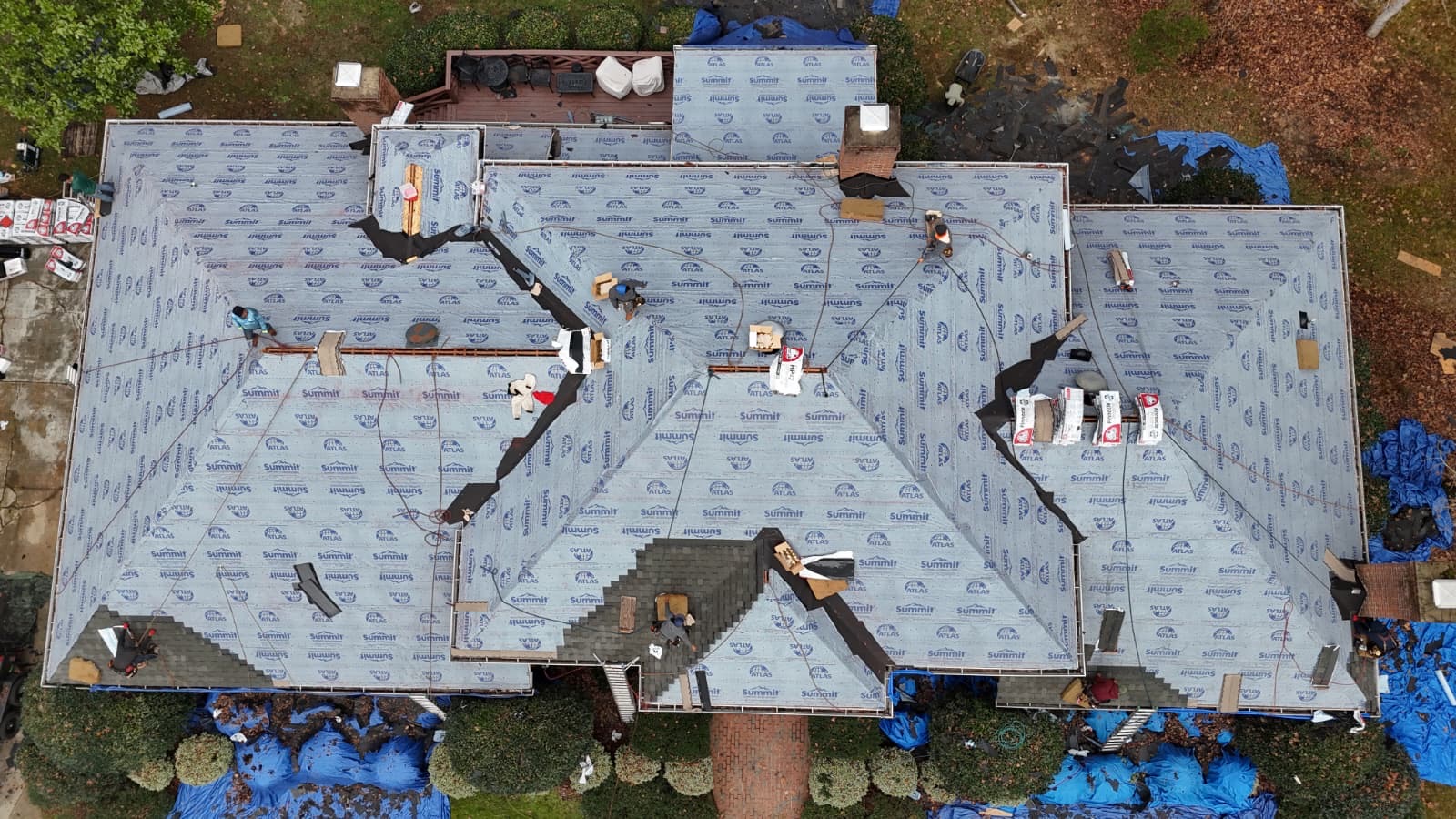 Aerial view of a Quail Hollow-style Charlotte estate home mid-installation, showing fresh Atlas Summit synthetic underlayment across a complex multi-gable hip roof with multiple chimneys, roofing crew at work, valley liners installed, and shingle bundles staged for the next phase