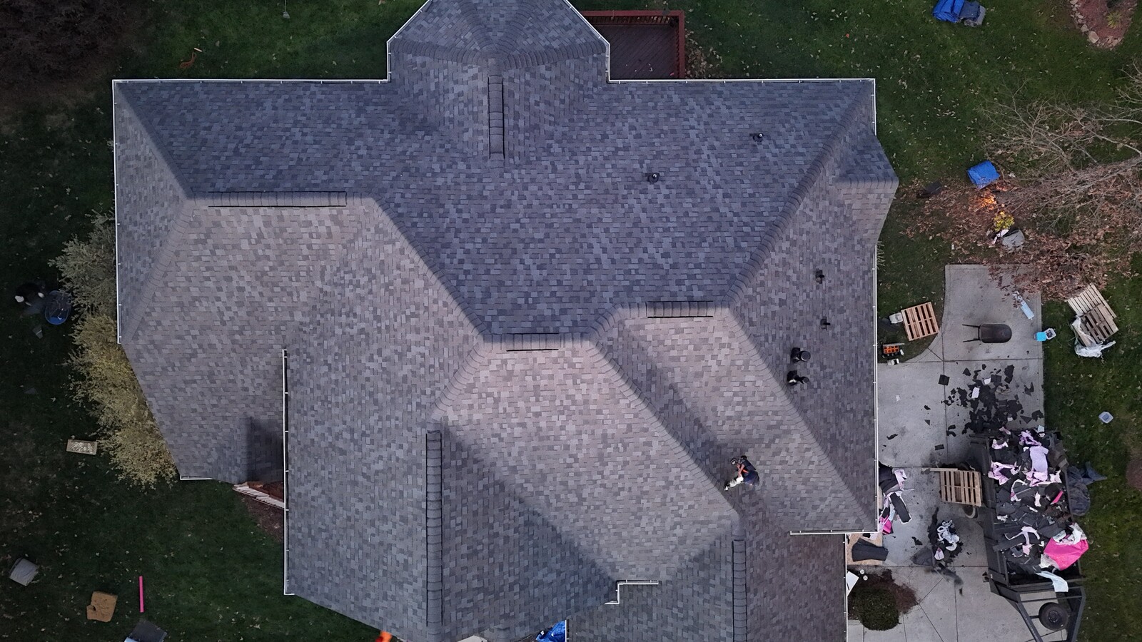 Aerial view of a completed gray and charcoal architectural-shingle roof on a large Quail Hollow-area estate home, showing crisp ridge caps on a complex multi-hip roofline with center gable peak, with Charlotte Ace Roofing crew finishing cleanup, dump trailer, and shingle waste palletized below