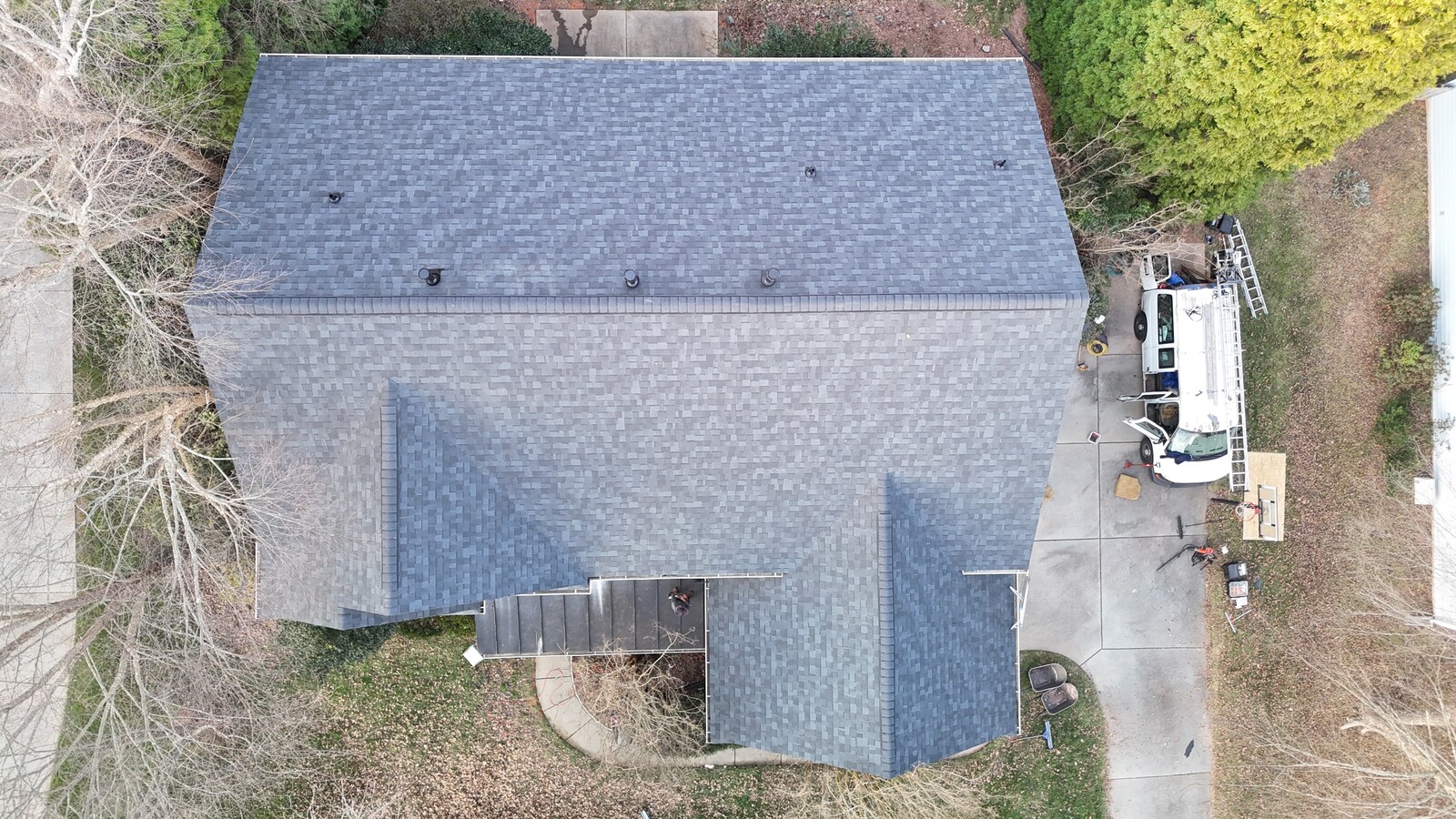 Aerial view of a completed Charlotte Ace Roofing project on a Sedgefield-area home with fresh charcoal-gray architectural shingles, a clean rectangular hip roof and small front-facing gable dormer, beige siding, with the contractor's white work van visible in the driveway