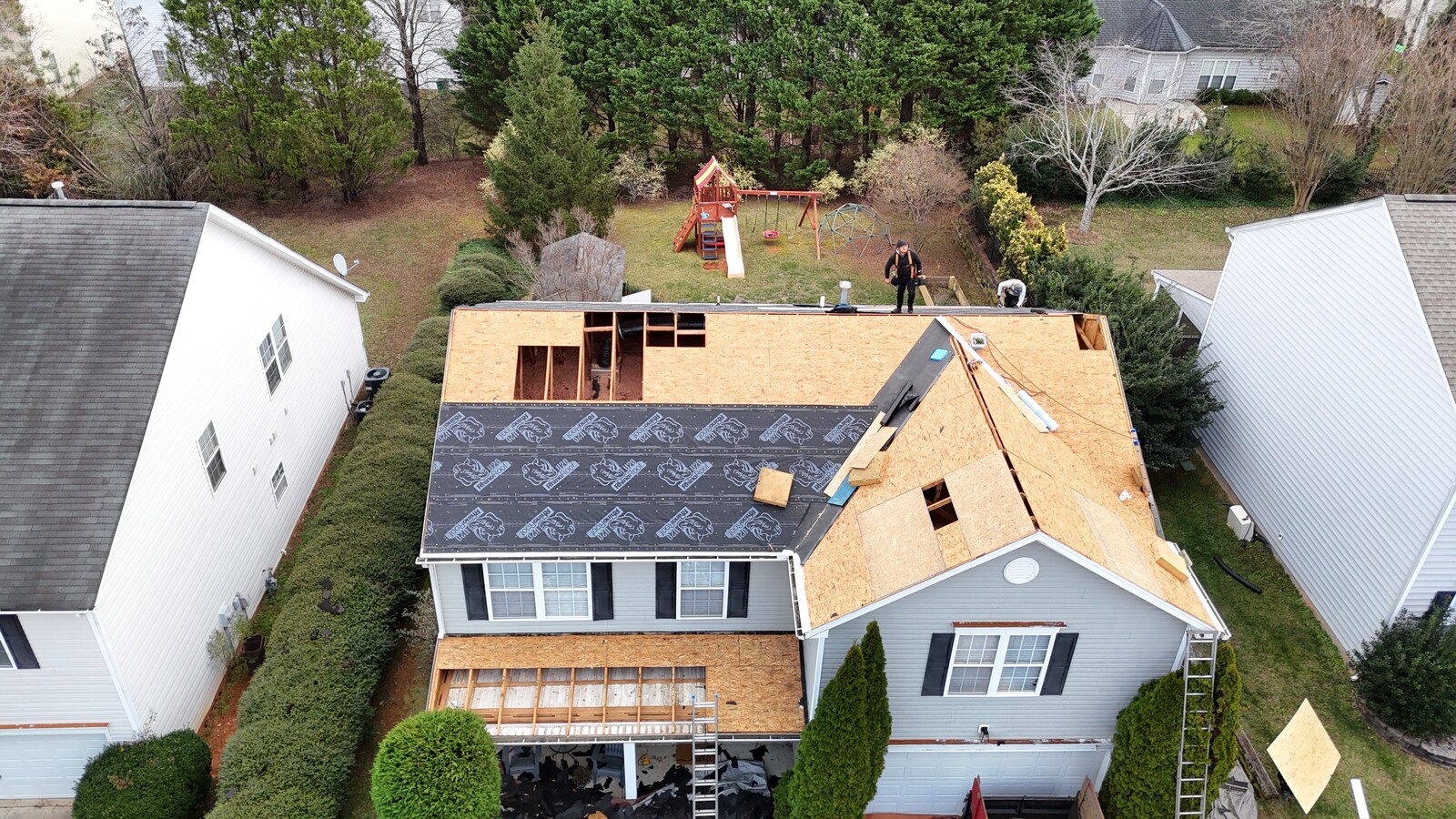 Aerial mid-installation view of a Sedgefield-area gray two-story Colonial home, showing the new OSB roof decking installed over the original 1940s framing with rectangular openings cut for skylights and ventilation, RhinoRoof U20 synthetic underlayment partially laid, two crew members visible installing materials, ladder set up at the front, and a backyard play set in the neighbor's yard