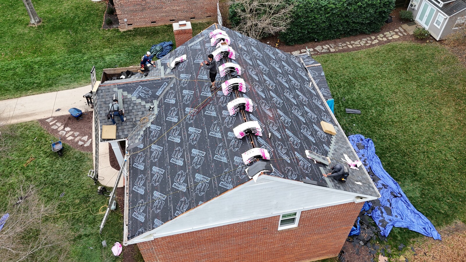 Aerial mid-installation view of a Sedgefield-area brick home with hip roof, RhinoRoof U20 synthetic underlayment fully installed, multiple bundles of Owens Corning Duration architectural shingles in pink wrap staged across the slope, four crew members actively working on the installation, dump tarp visible at the side with old shingle debris, and a wheelbarrow staged on the front walk