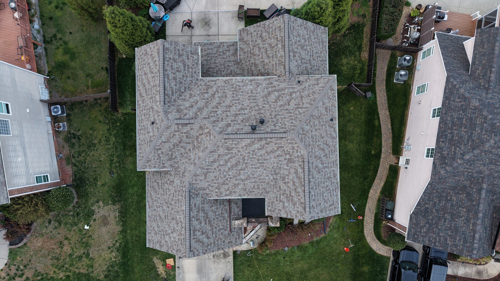 Aerial view of a Charlotte Ace Roofing project on a complex multi-gable suburban home with a freshly installed gray and tan blend architectural-shingle roof, showing crisp ridge-cap detailing across the multiple roof intersections, a rear concrete patio area, neighboring homes on either side, and a paver walkway leading to the back of the property