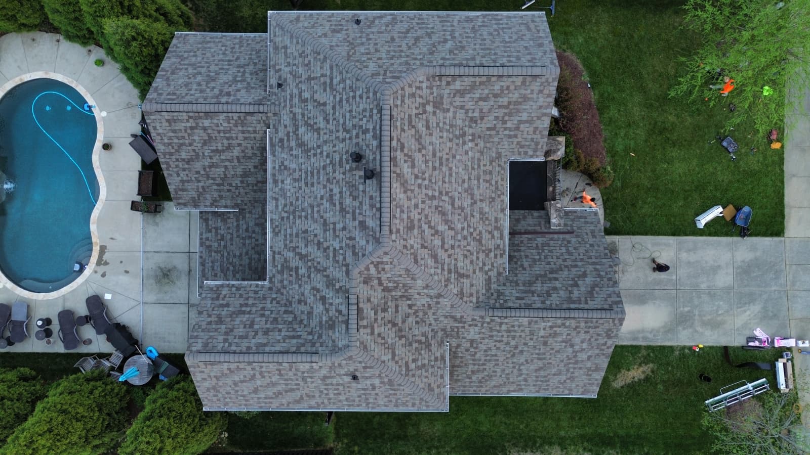 Aerial view of a completed Charlotte Ace Roofing project on a luxury home with a kidney-shaped pool and pool deck visible to the left, showing a fresh tan and brown blend architectural-shingle roof on a complex multi-hip and multi-gable roofline, with concrete pool patio, lounge chairs, and crew member visible on the side driveway