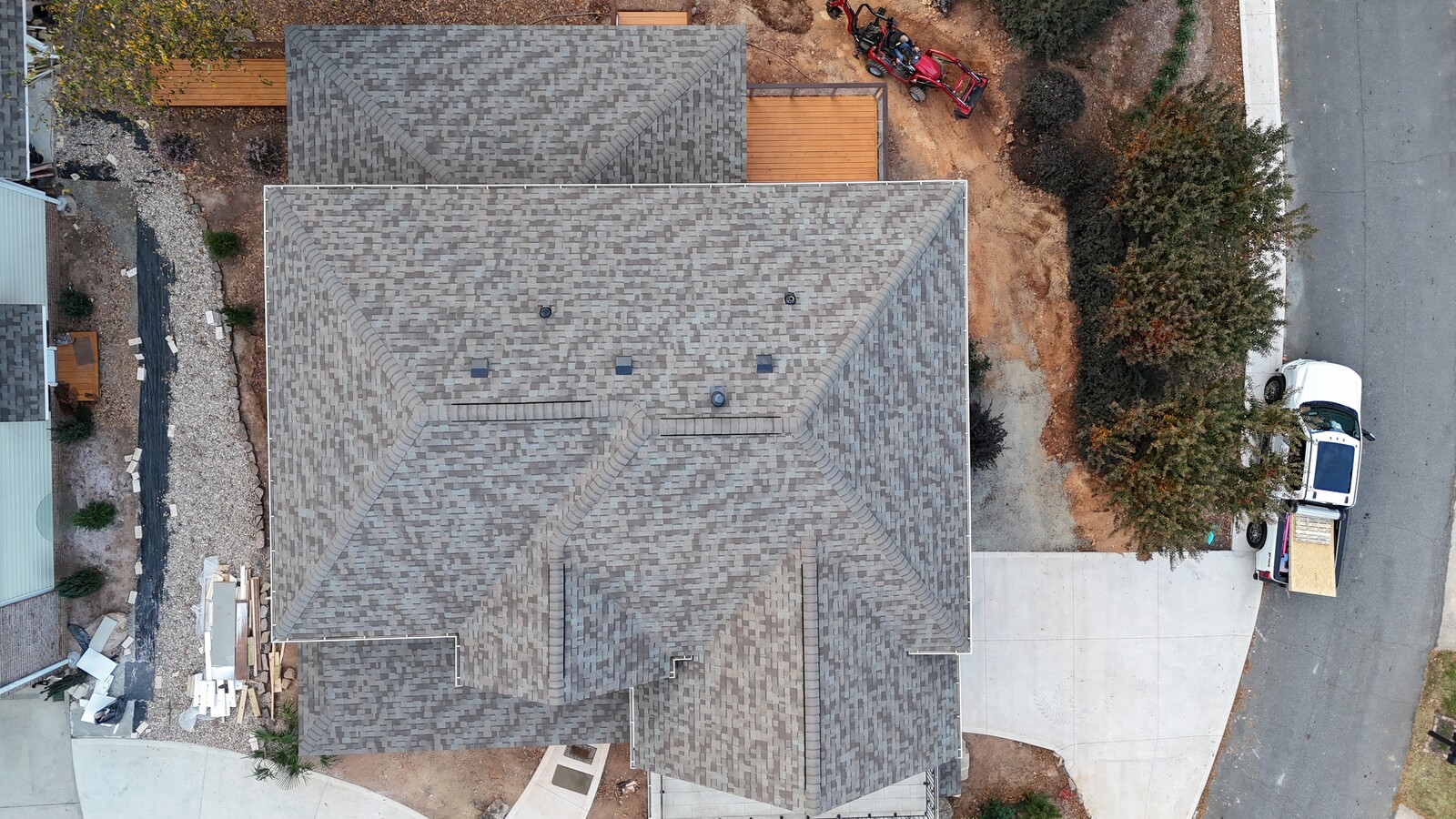 Aerial view of a completed Charlotte Ace Roofing project on a large home and detached structure, showing fresh gray and charcoal blend architectural shingles on both buildings, crisp hip-and-gable ridge detailing, the contractor's white work van and equipment trailer parked in the driveway, and active landscape work in progress in the front yard
