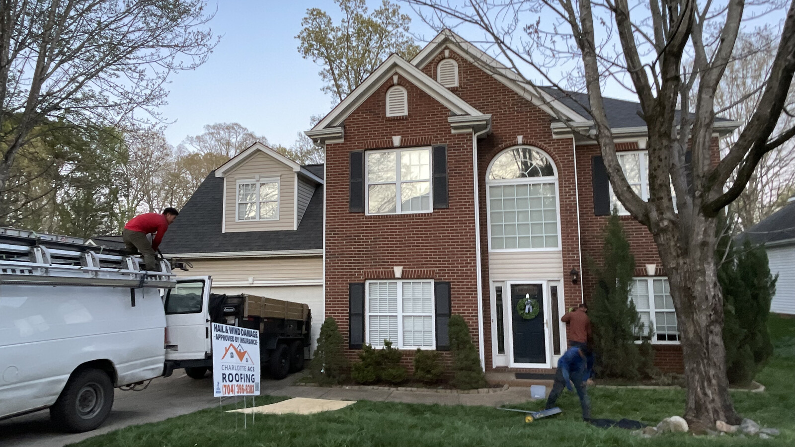 Charlotte Ace Roofing crew on the roof of a two-story brick home in Stallings, NC during a hail-damage roof replacement — company van and dump trailer in driveway, yard sign visible