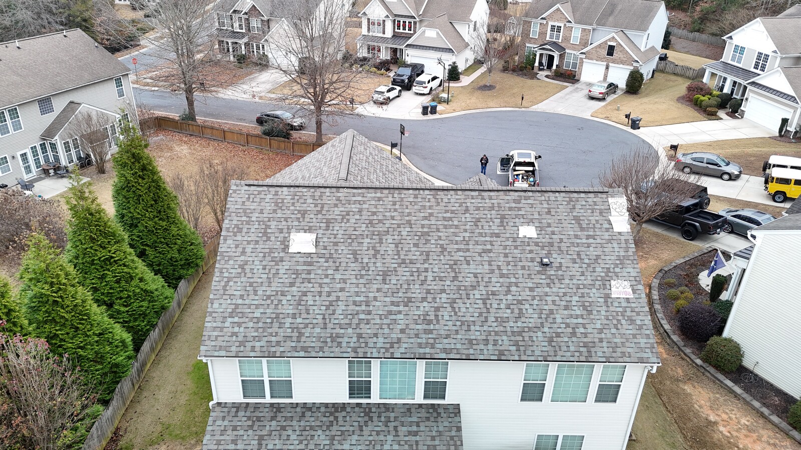 Aerial view of a University City-area subdivision home with a freshly installed gray architectural-shingle roof, showing the contractor's white work van parked at the cul-de-sac with crew member ready to work, neighboring 2-story brick-and-vinyl homes with mature landscaping, and the suburban subdivision context typical of the U-City area
