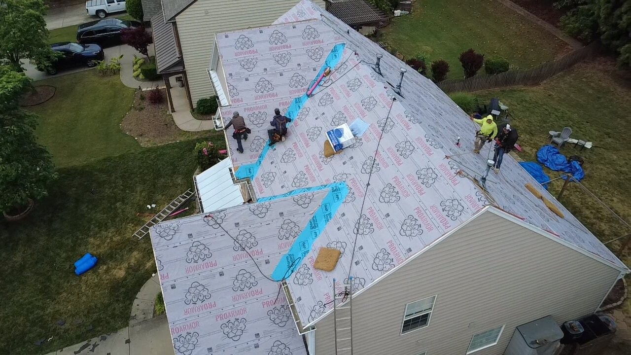 Aerial mid-installation view of a University City-area home showing fresh ProArmor synthetic underlayment fully installed across the entire roof slope with chalk-line layout marks for shingle courses, four crew members actively working including a man in a yellow safety vest, ladders set against the front of the home, and tarps protecting landscaping below