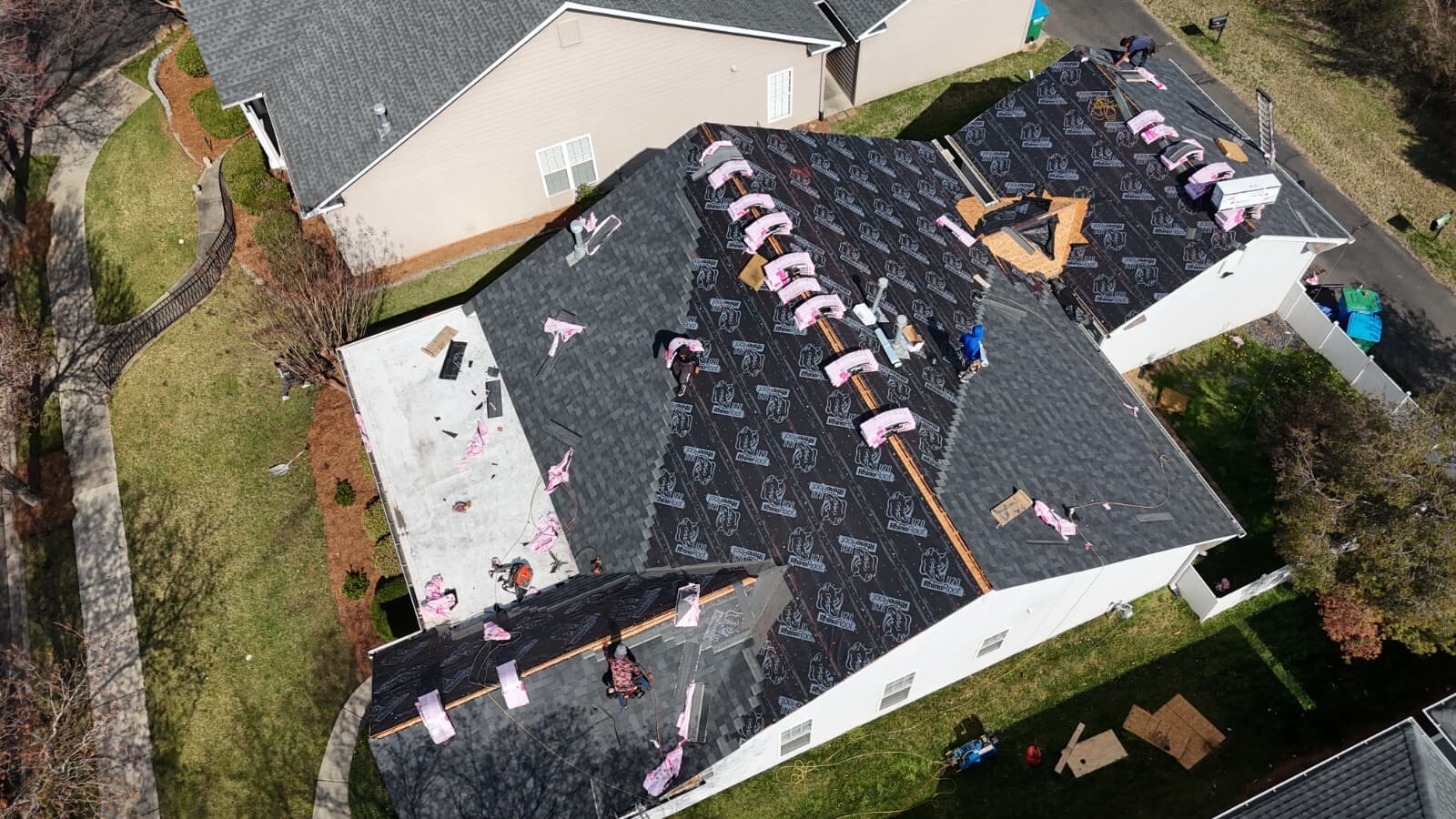 Aerial view of a University City-area subdivision home mid-installation, showing partially completed dark gray architectural shingles starting from the lower roof edge with RhinoRoof U20 synthetic underlayment laid across the upper sections, multiple bundles of Owens Corning Duration shingles in pink wrap staged for next-phase installation across the slope, and crew members actively working on the front of the property