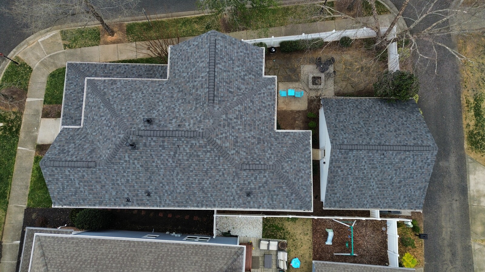 Aerial view of an Uptown-area home with a freshly installed gray and charcoal blend architectural-shingle roof on a complex multi-gable roofline with prominent center peak, showing crisp ridge cap detailing on every slope intersection, the home situated on a tight urban lot with mature trees, neighboring older Charlotte homes visible, and a stone patio area at the rear of the property