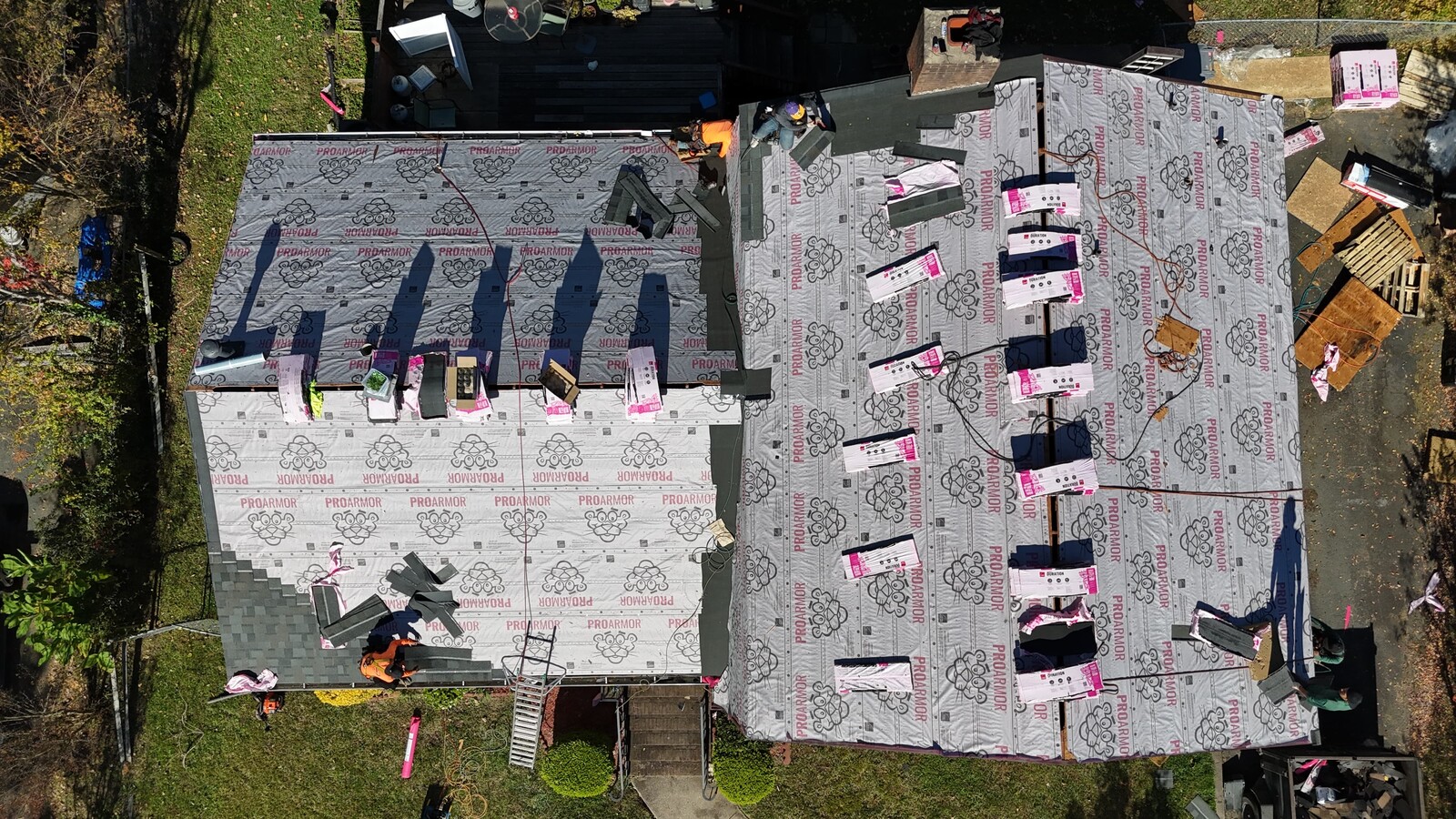 Aerial mid-installation view of an older multi-section Uptown-area home, showing fresh ProArmor synthetic underlayment laid across both wings of the property with bundles of Owens Corning Duration shingles staged for installation, multiple crew members actively working on different sections including one on the back deck area near the brick chimney, and a back yard with deck and grill visible