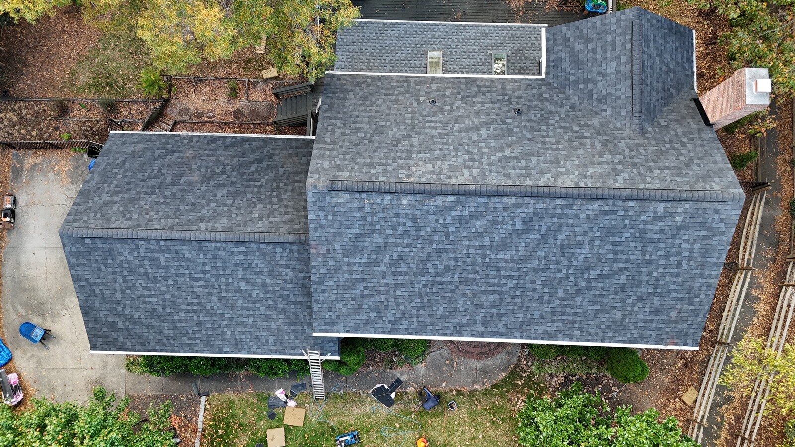Aerial view of a completed Uptown-area home with a multi-section roof showing fresh dark gray architectural shingles, including a front lower extension and a taller rear two-story section with brick chimney, surrounded by mature wooded landscaping with autumn-colored trees, and a brick walkway and steps leading to the back of the property